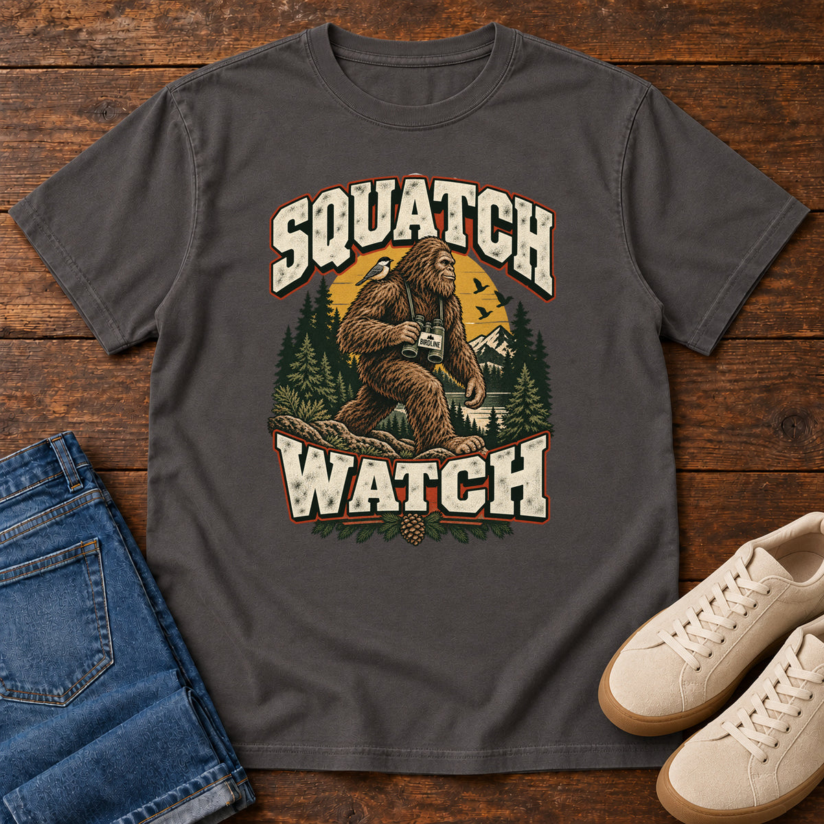 Squatch Watch T-Shirt in Graphite, front laid flat on wood, featuring a walking Sasquatch holding binoculars among pine trees, framed by large cream lettering.