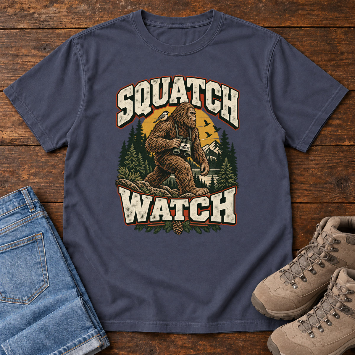 Squatch Watch T-Shirt in Navy, front laid flat on wood, featuring a walking Sasquatch holding binoculars among pine trees, framed by large cream lettering.