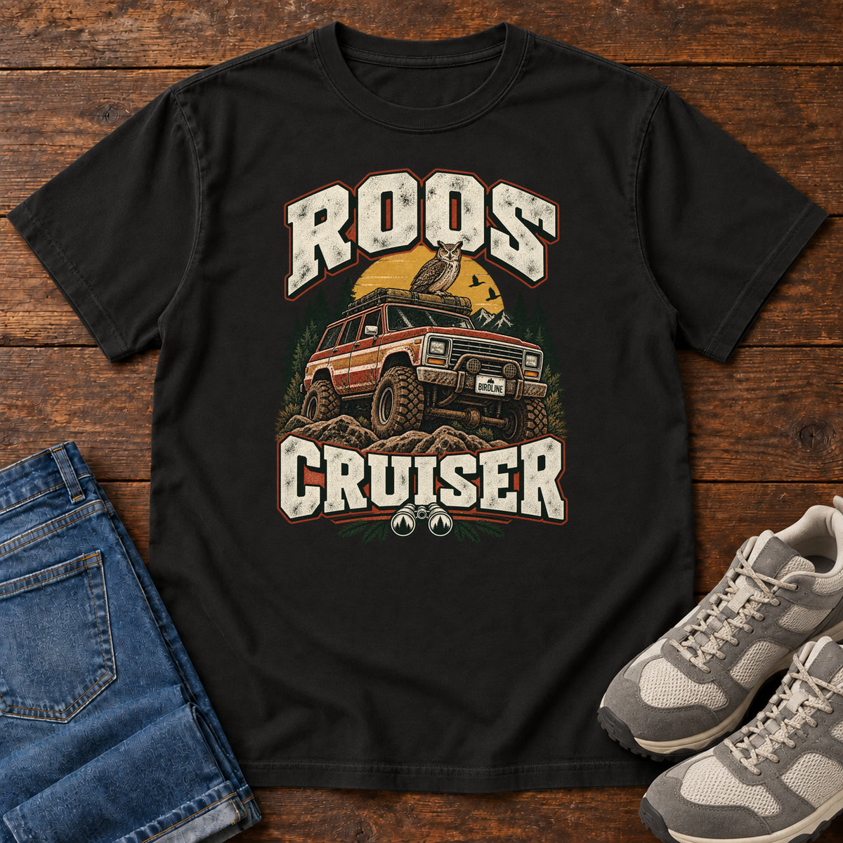 Black Roos Cruiser T-Shirt with a bird perched on an off-road vehicle between distressed cream Roos Cruiser lettering, front flat lay on wood.