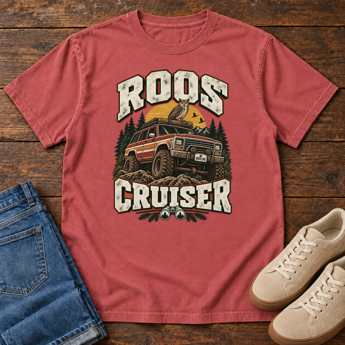 Crimson Roos Cruiser T-Shirt with a bird perched on an off-road vehicle between distressed cream Roos Cruiser lettering, front flat lay on wood.