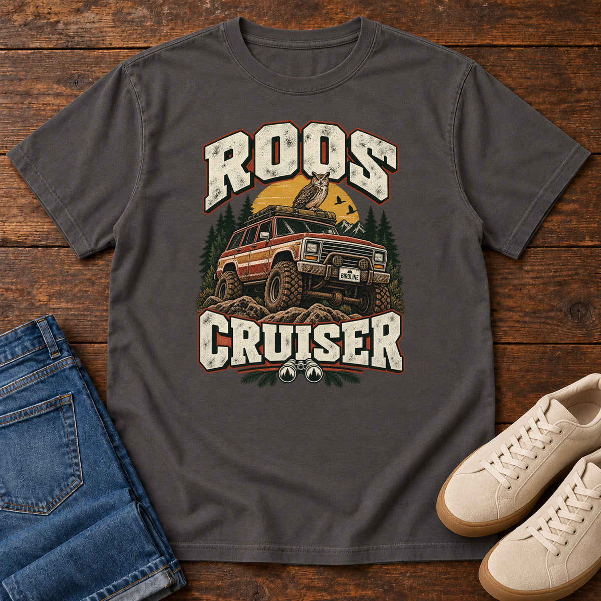 Graphite Roos Cruiser T-Shirt with a bird perched on an off-road vehicle between distressed cream Roos Cruiser lettering, front flat lay on wood.