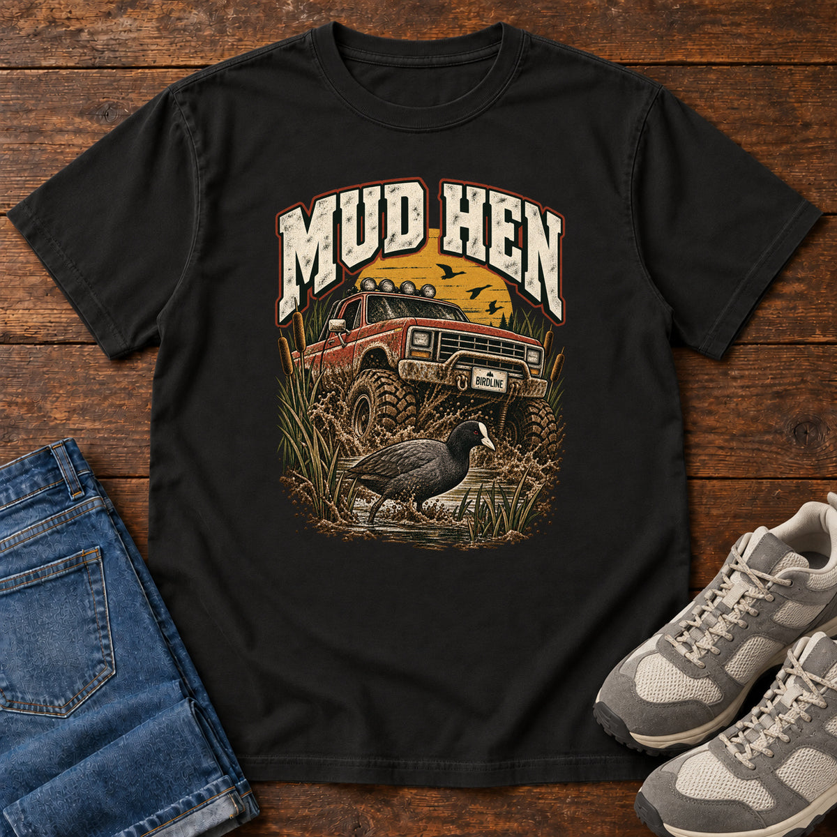Black Mud Hen T-Shirt with a red off-road truck splashing through mud behind a dark waterbird and reeds; front flat lay on wood.