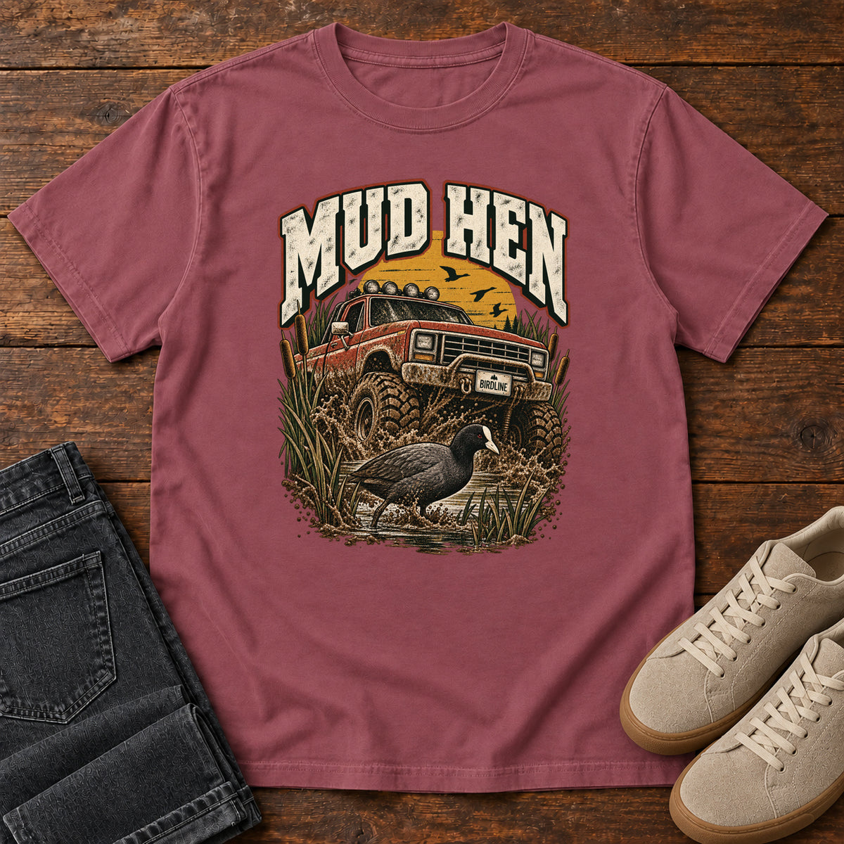 Brick Mud Hen T-Shirt with a red off-road truck splashing through mud behind a dark waterbird and reeds; front flat lay on wood.