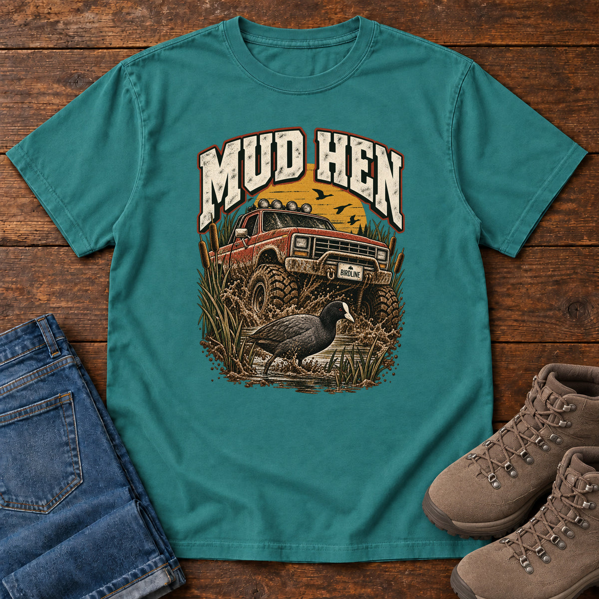 Emerald Mud Hen T-Shirt with a red off-road truck splashing through mud behind a dark waterbird and reeds; front flat lay on wood.