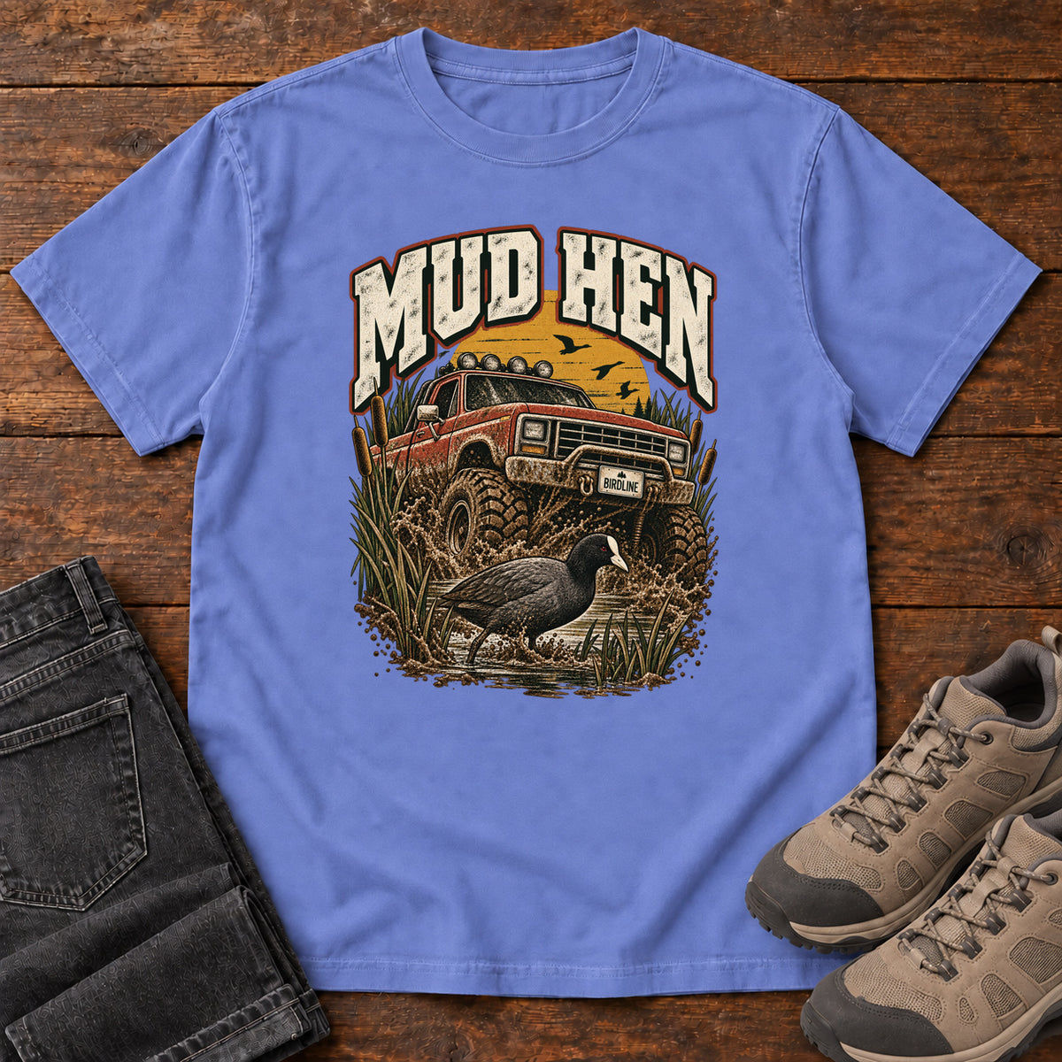 Flo Blue Mud Hen T-Shirt with a red off-road truck splashing through mud behind a dark waterbird and reeds; front flat lay on wood.