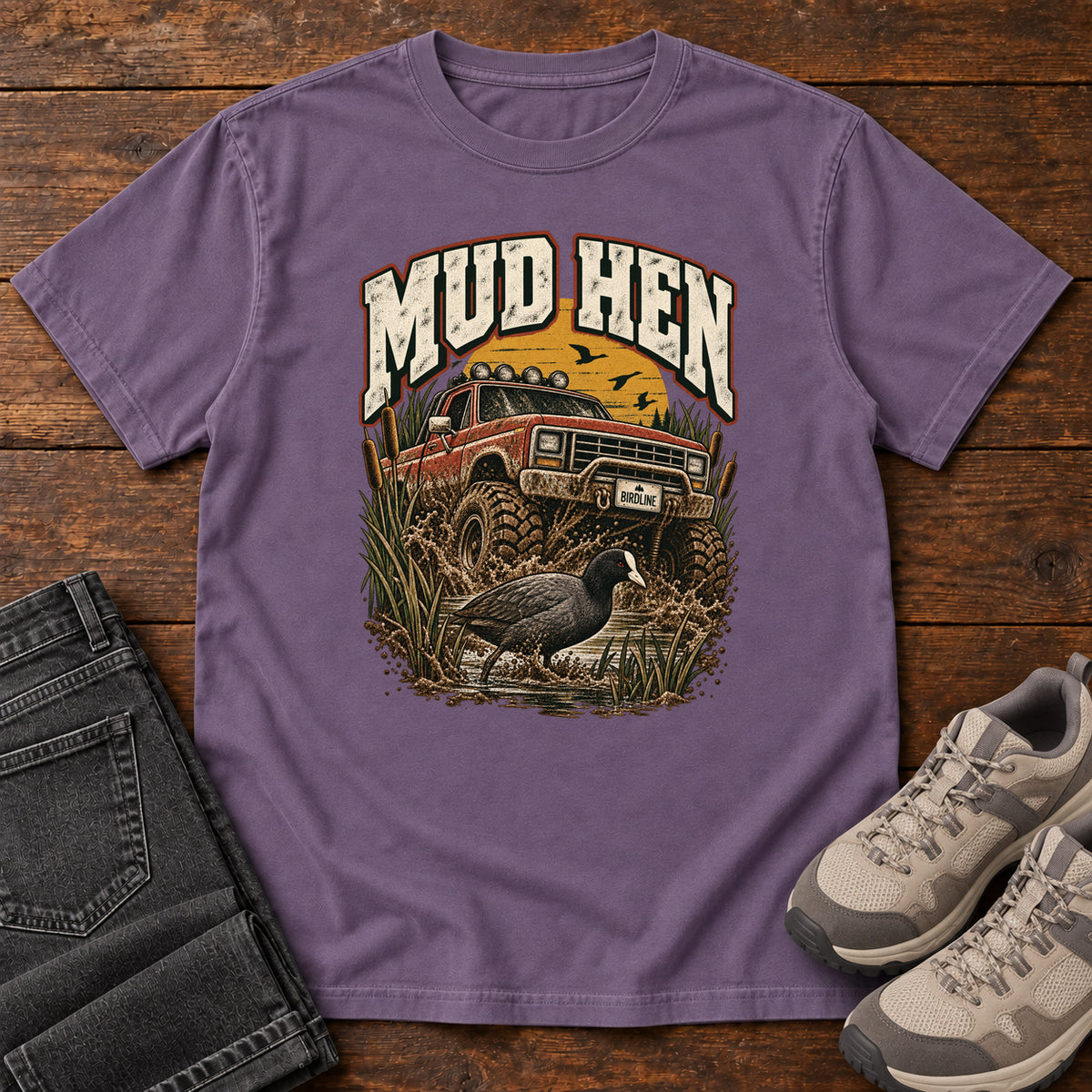 Grape Mud Hen T-Shirt with a red off-road truck splashing through mud behind a dark waterbird and reeds; front flat lay on wood.