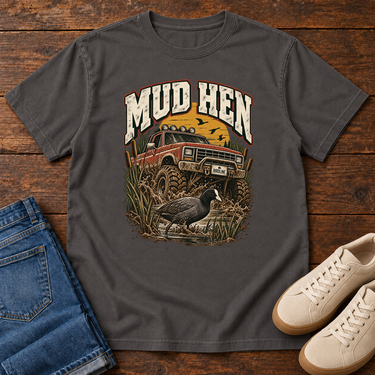 Graphite Mud Hen T-Shirt with a red off-road truck splashing through mud behind a dark waterbird and reeds; front flat lay on wood.