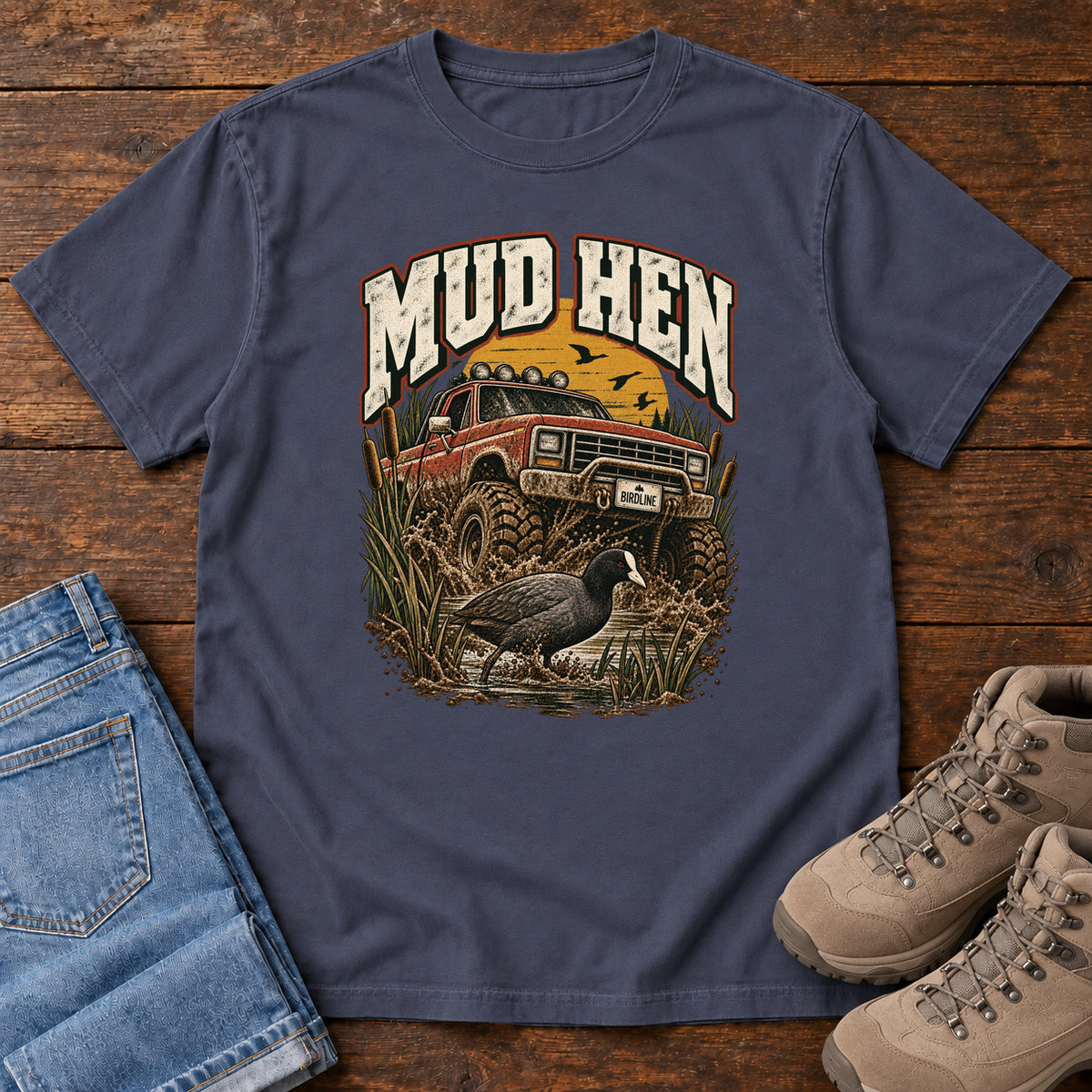 Navy Mud Hen T-Shirt with a red off-road truck splashing through mud behind a dark waterbird and reeds; front flat lay on wood.