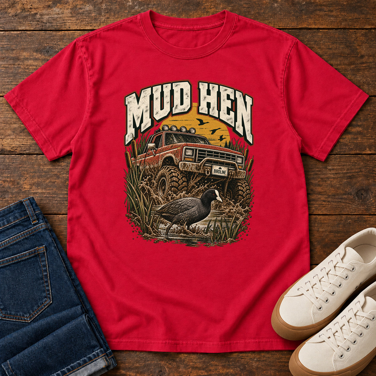 Red Mud Hen T-Shirt with a red off-road truck splashing through mud behind a dark waterbird and reeds; front flat lay on wood.