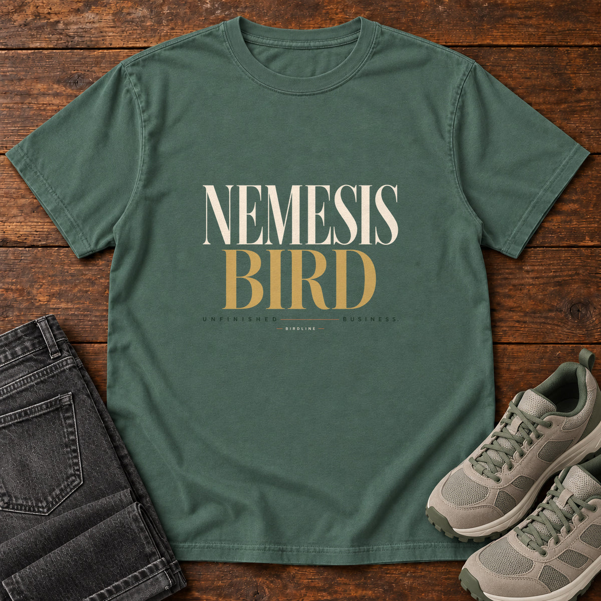 Blue Spruce Nemesis Bird T-Shirt with cream-and-gold serif lettering, front flat lay on wood.