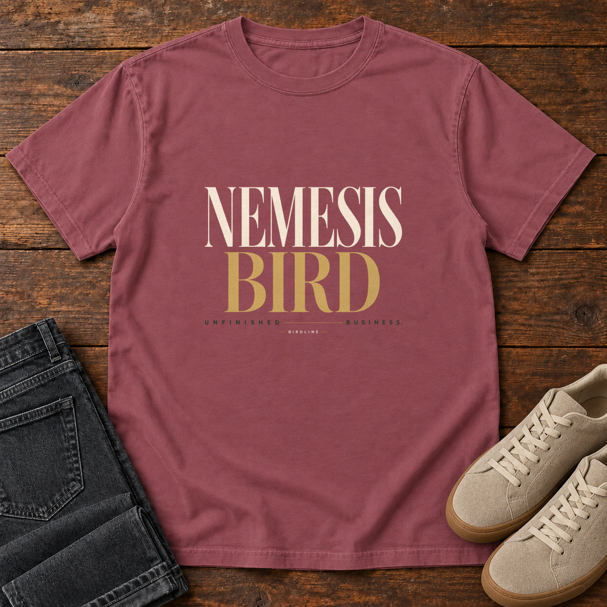 Brick Nemesis Bird T-Shirt with cream-and-gold serif lettering, front flat lay on wood.