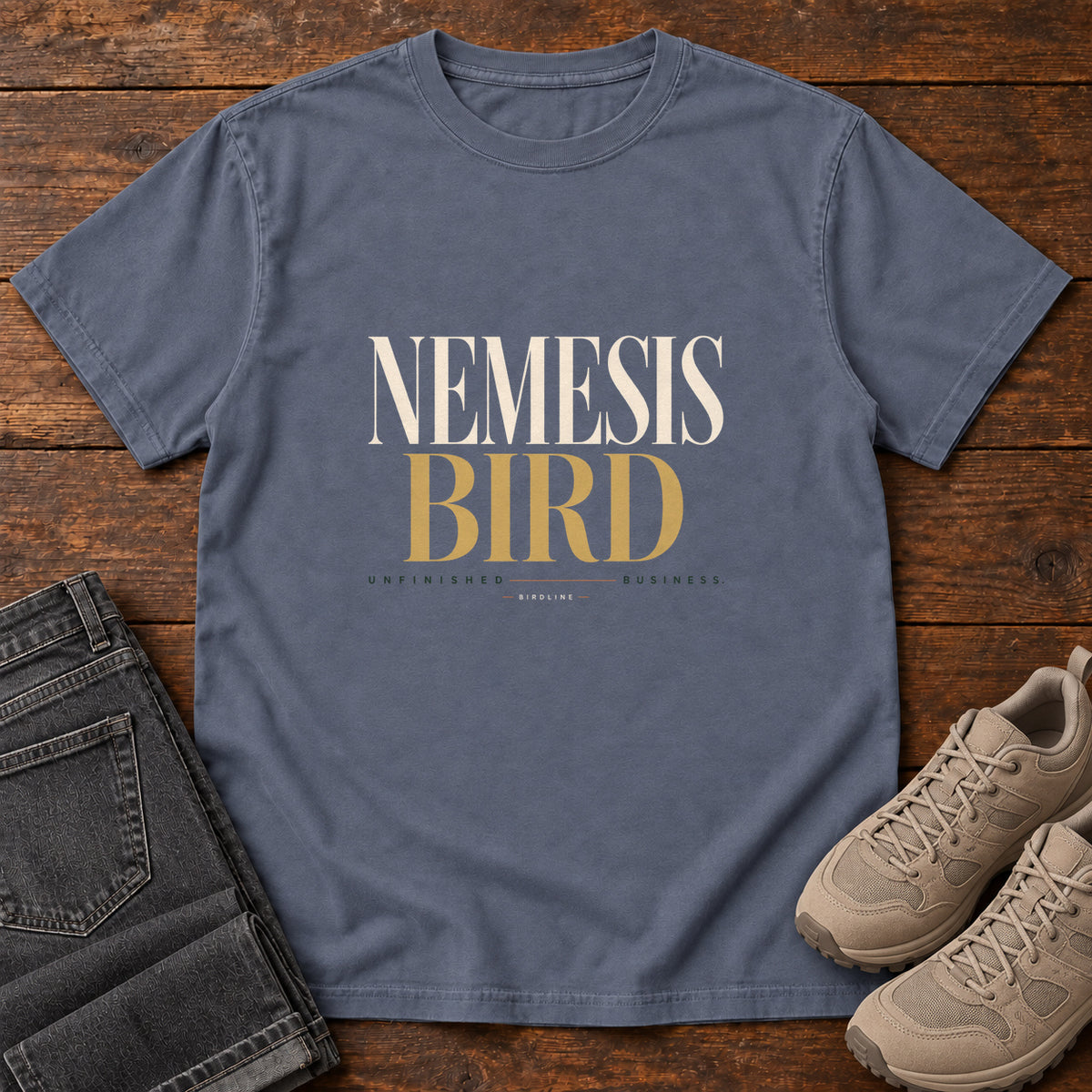 Denim Nemesis Bird T-Shirt with cream-and-gold serif lettering, front flat lay on wood.
