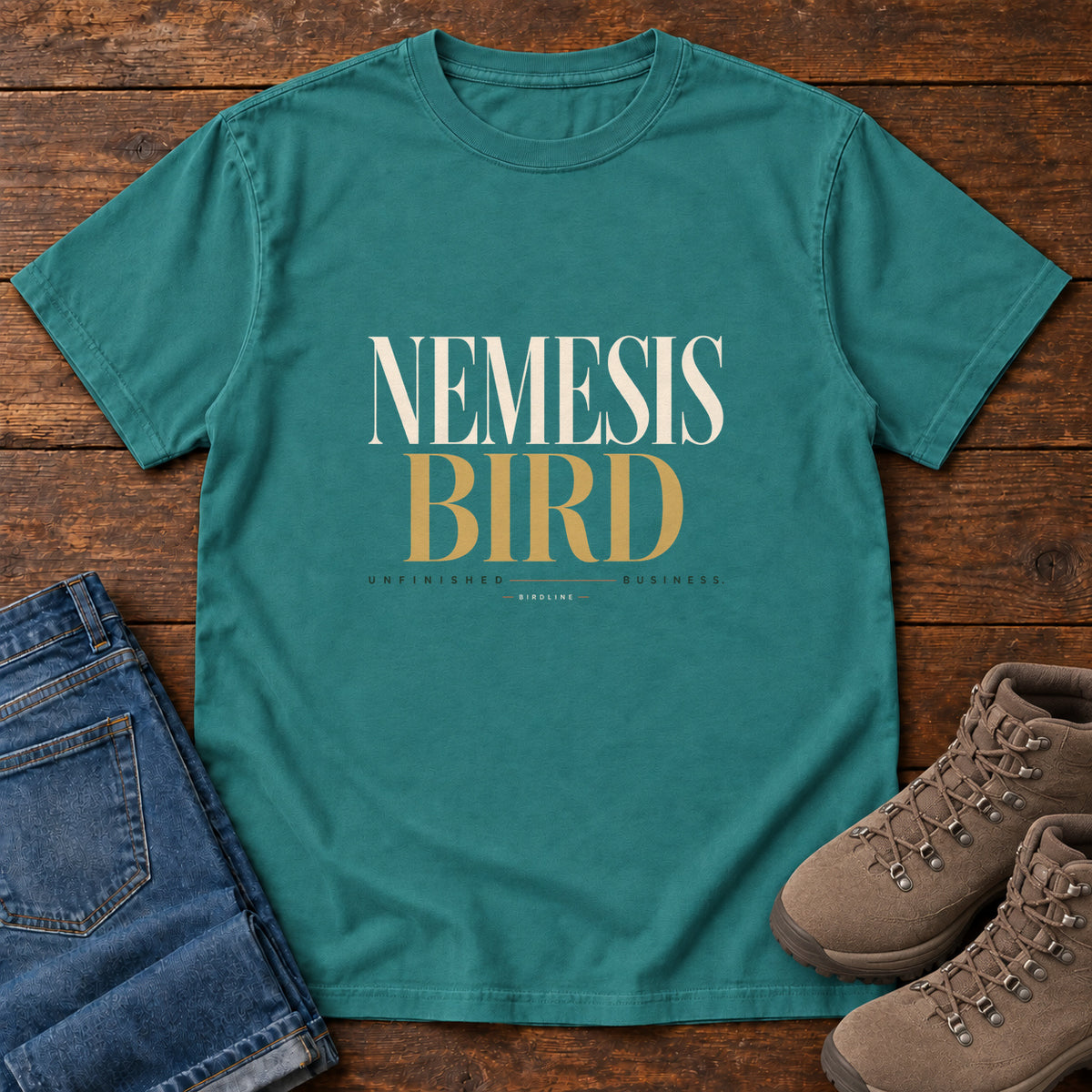 Emerald Nemesis Bird T-Shirt with cream-and-gold serif lettering, front flat lay on wood.