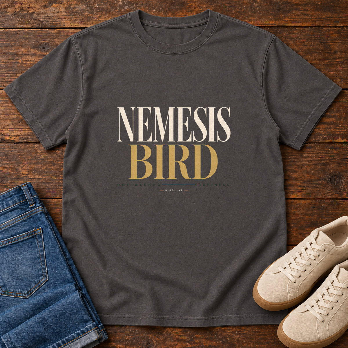 Graphite Nemesis Bird T-Shirt with cream-and-gold serif lettering, front flat lay on wood.