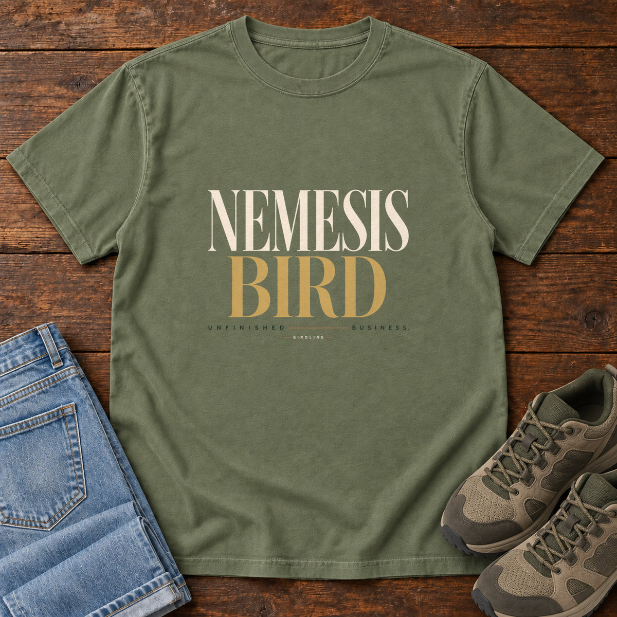 Moss Nemesis Bird T-Shirt with cream-and-gold serif lettering, front flat lay on wood.