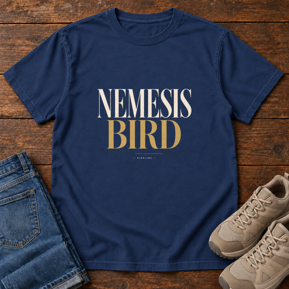 True Navy Nemesis Bird T-Shirt with cream-and-gold serif lettering, front flat lay on wood.