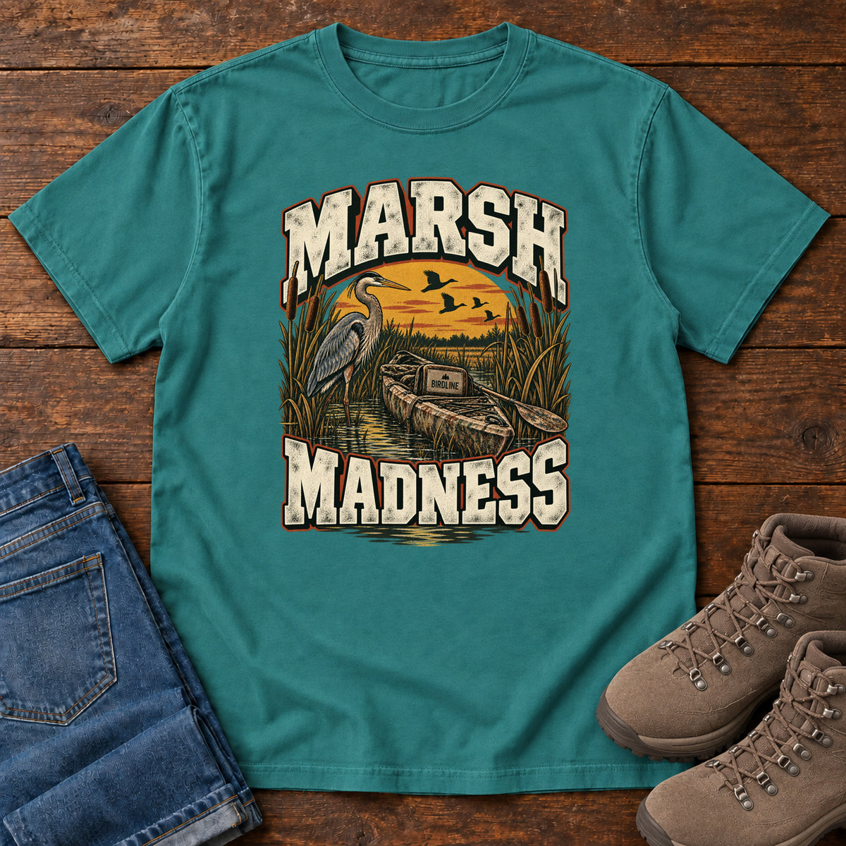 Emerald Marsh Madness T-Shirt with a heron and canoe among marsh reeds beneath an orange sky and flying birds; front flat lay on wood.