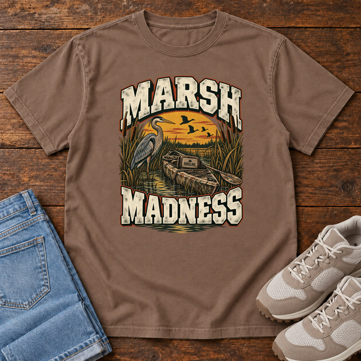 Espresso Marsh Madness T-Shirt with a heron and canoe among marsh reeds beneath an orange sky and flying birds; front flat lay on wood.