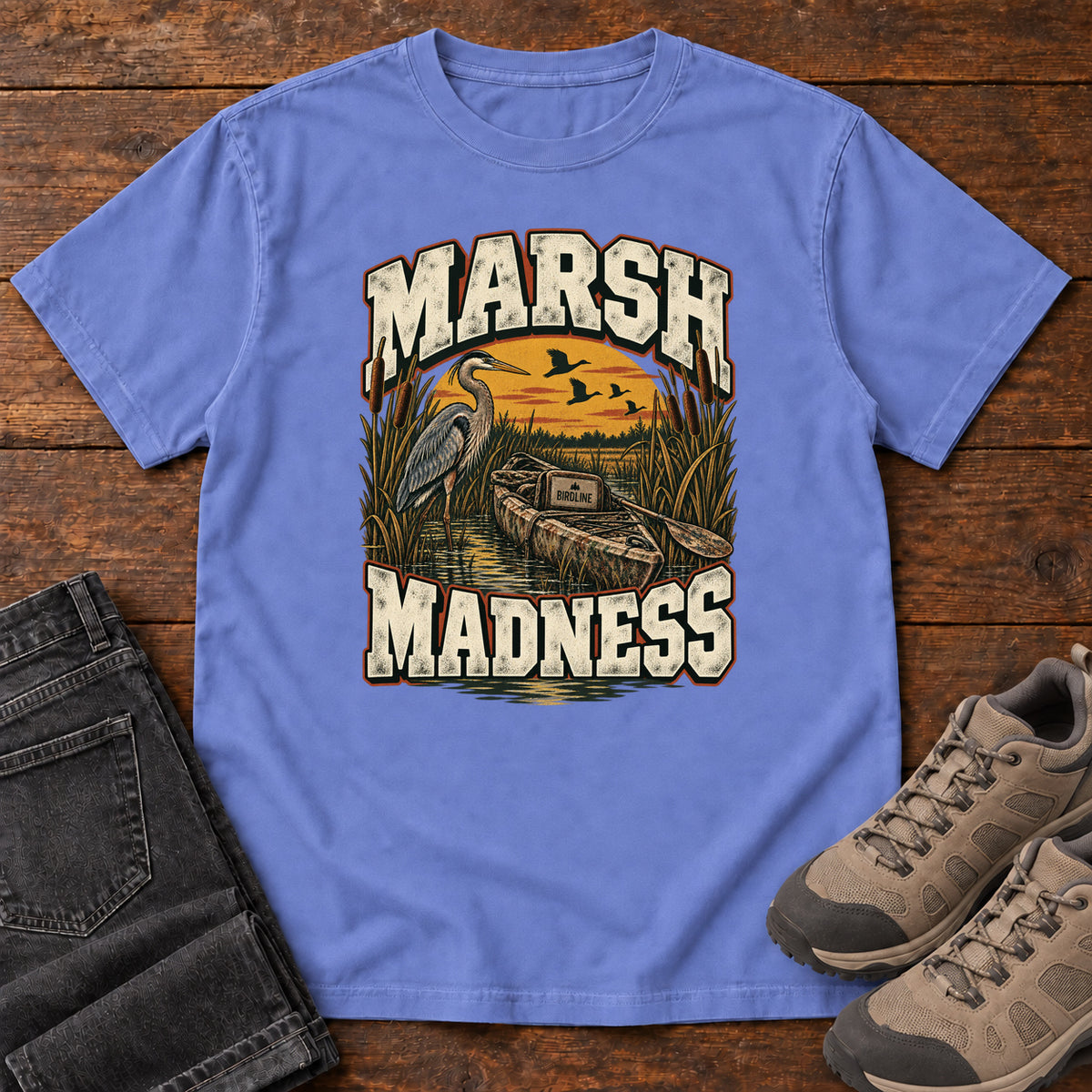 Flo Blue Marsh Madness T-Shirt with a heron and canoe among marsh reeds beneath an orange sky and flying birds; front flat lay on wood.
