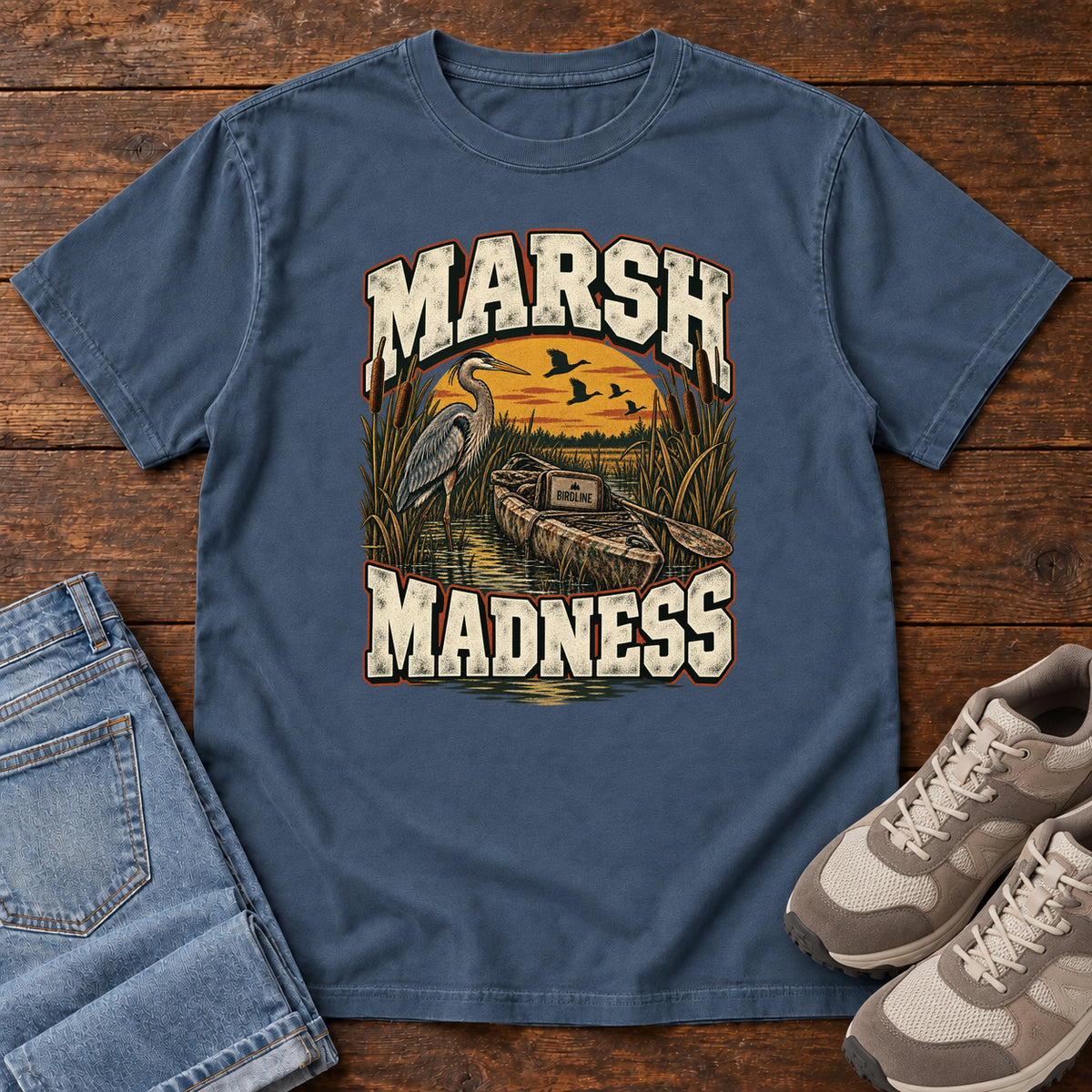 Midnight Marsh Madness T-Shirt with a heron and canoe among marsh reeds beneath an orange sky and flying birds; front flat lay on wood.