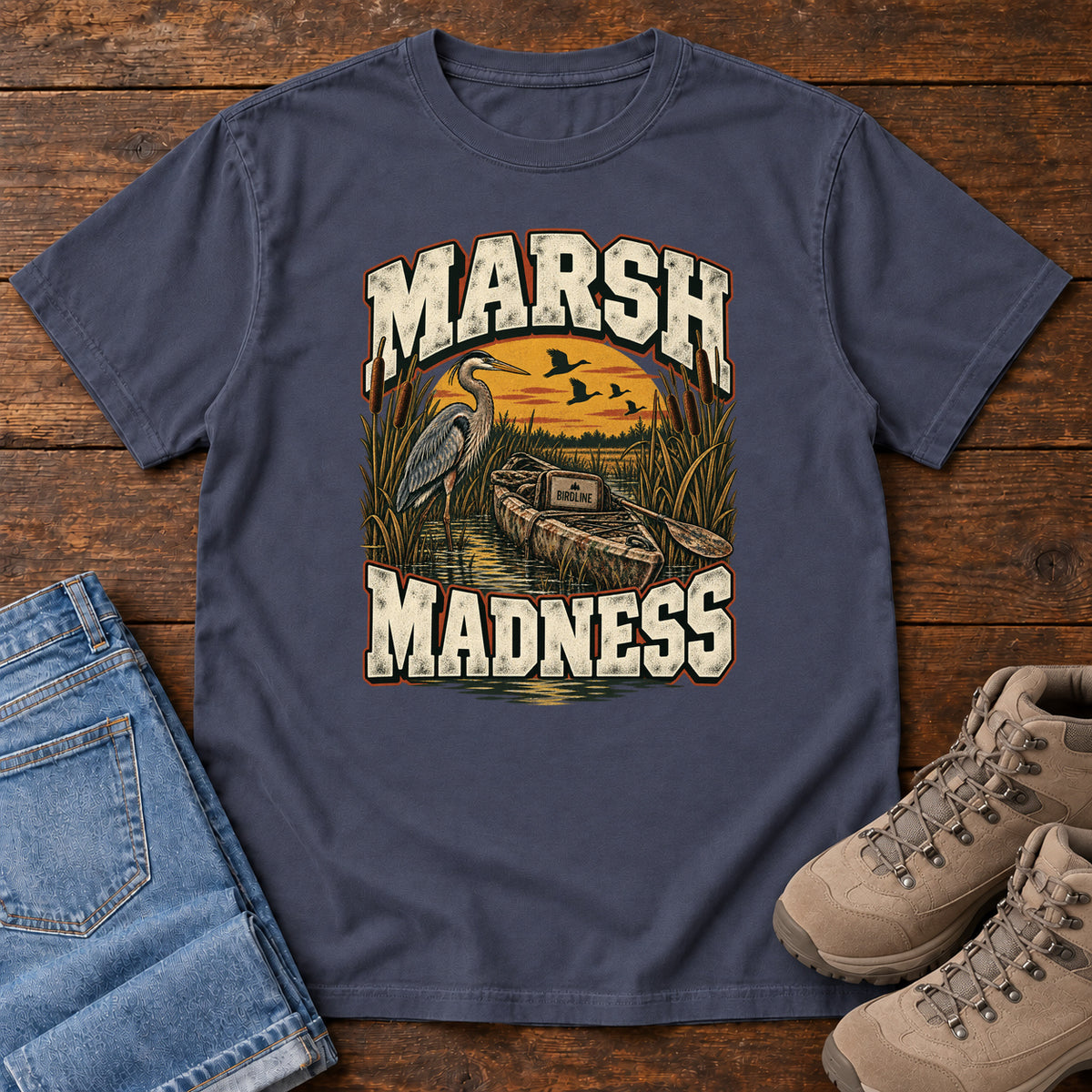Navy Marsh Madness T-Shirt with a heron and canoe among marsh reeds beneath an orange sky and flying birds; front flat lay on wood.