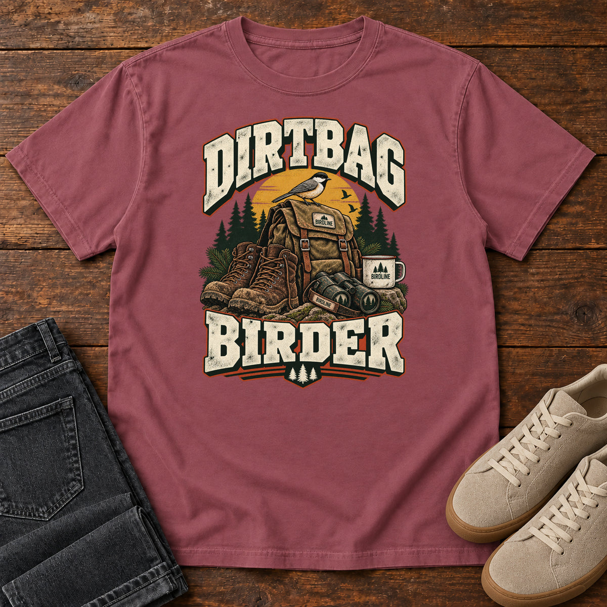 Brick Dirtbag Birder T-Shirt with hiking boots, a backpack, binoculars and a camping mug beneath cream lettering, front flat lay on wood.
