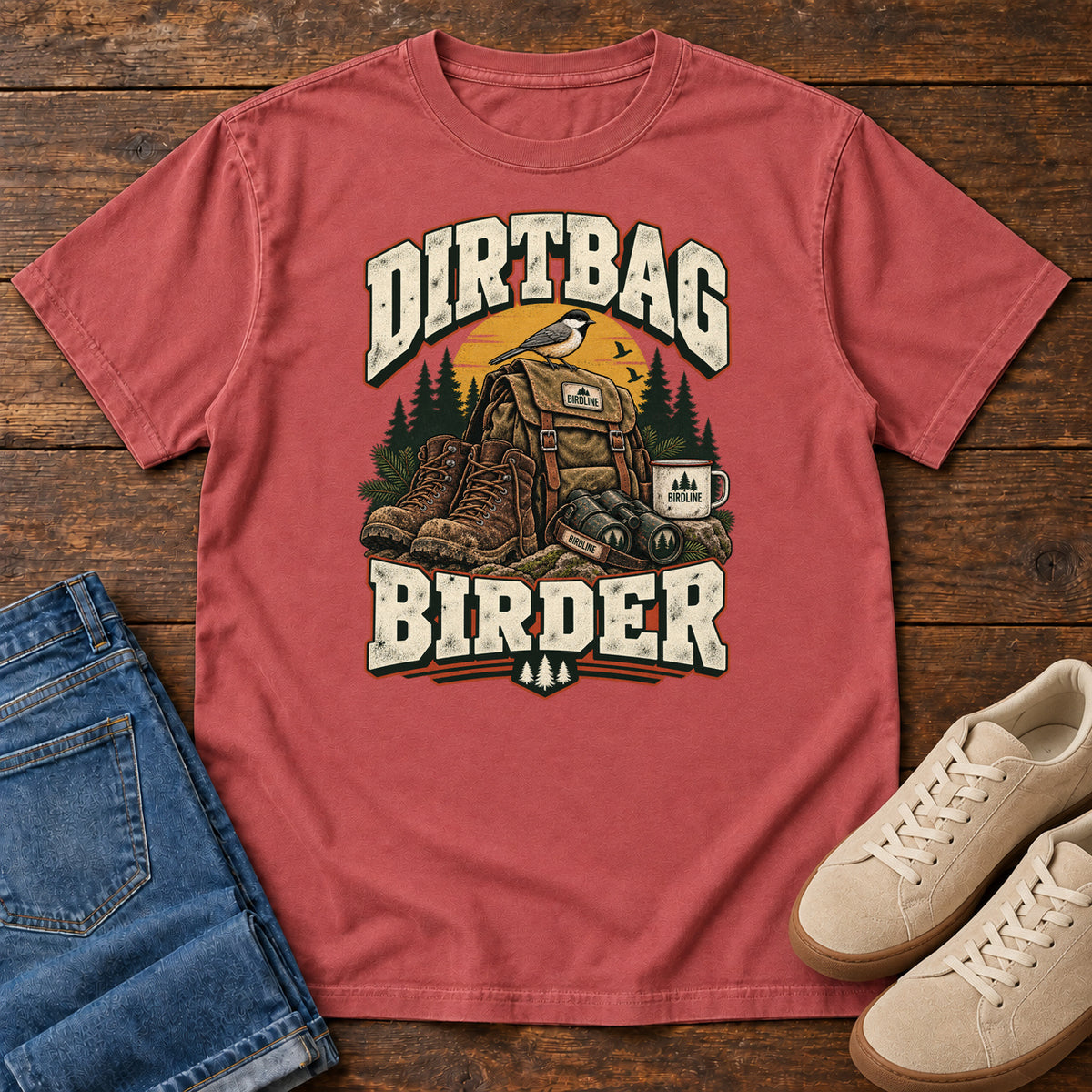 Crimson Dirtbag Birder T-Shirt with hiking boots, a backpack, binoculars and a camping mug beneath cream lettering, front flat lay on wood.