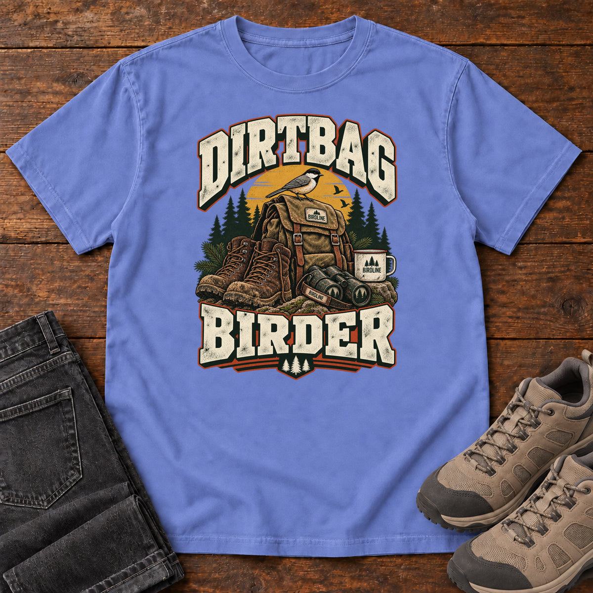 Flo Blue Dirtbag Birder T-Shirt with hiking boots, a backpack, binoculars and a camping mug beneath cream lettering, front flat lay on wood.