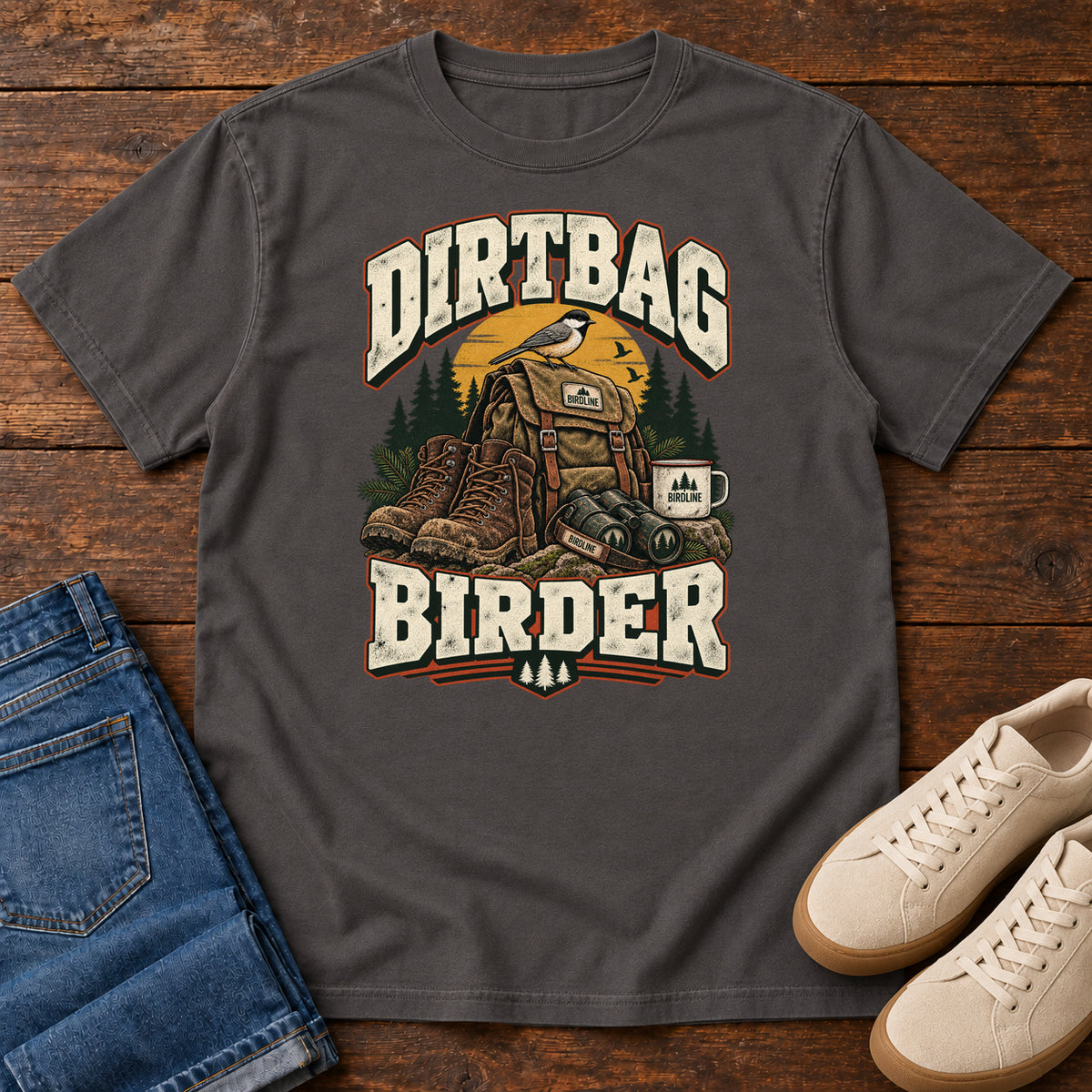 Graphite Dirtbag Birder T-Shirt with hiking boots, a backpack, binoculars and a camping mug beneath cream lettering, front flat lay on wood.