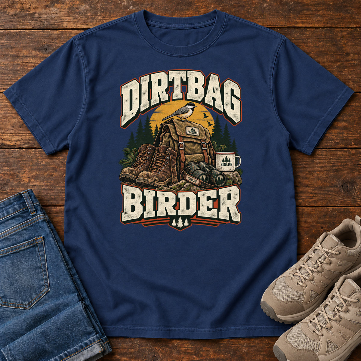 True Navy Dirtbag Birder T-Shirt with hiking boots, a backpack, binoculars and a camping mug beneath cream lettering, front flat lay on wood.