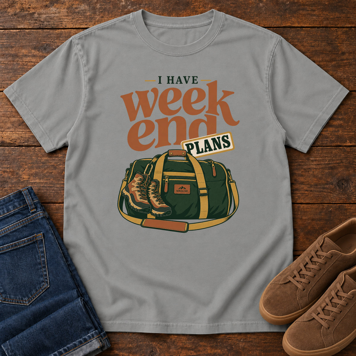 Grey I Have Weekend Plans T-Shirt with a green duffel bag and hiking boots beneath rust-orange Weekend lettering, front flat lay on wood.