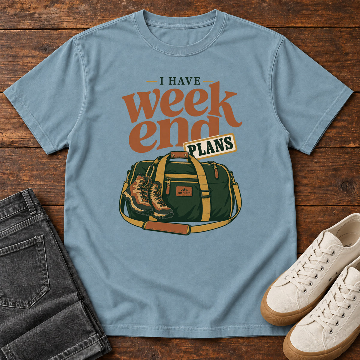 Ice Blue I Have Weekend Plans T-Shirt with a green duffel bag and hiking boots beneath rust-orange Weekend lettering, front flat lay on wood.