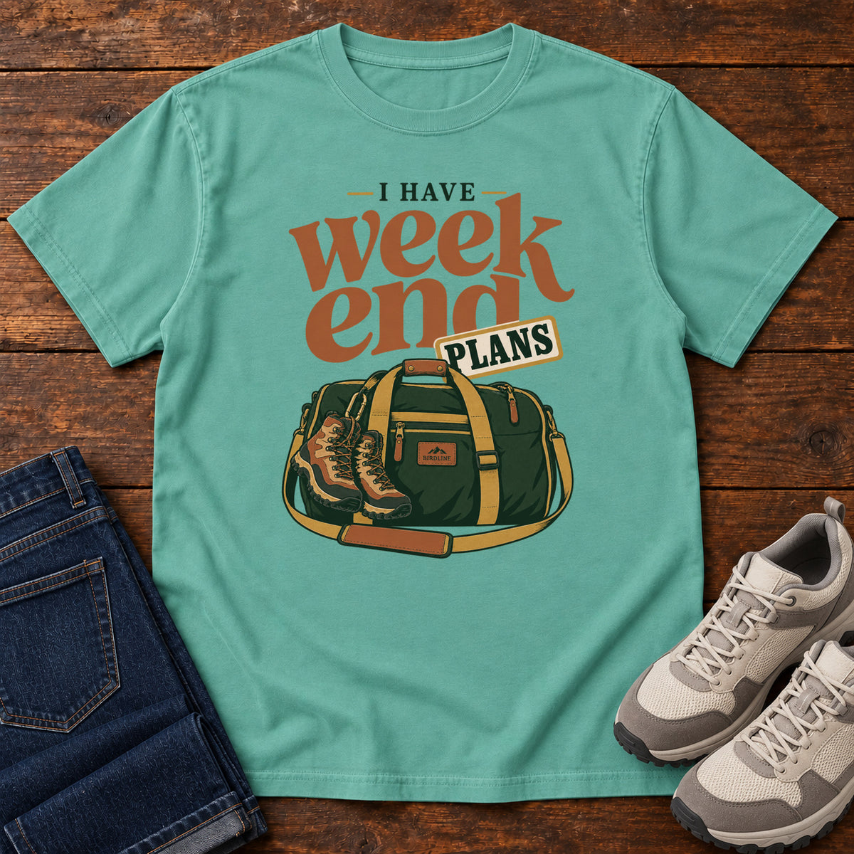 Seafoam I Have Weekend Plans T-Shirt with a green duffel bag and hiking boots beneath rust-orange Weekend lettering, front flat lay on wood.