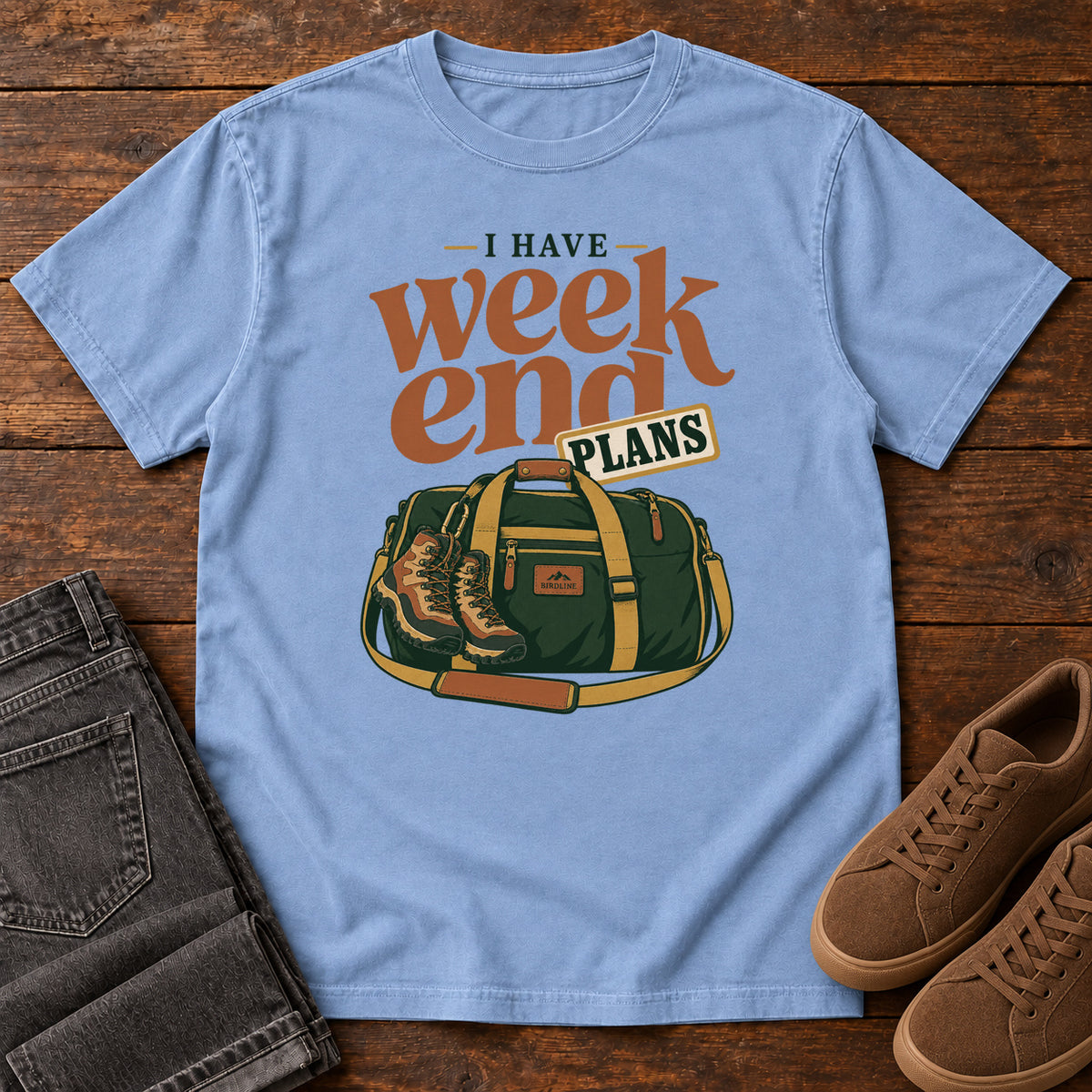 Washed Denim I Have Weekend Plans T-Shirt with a green duffel bag and hiking boots beneath rust-orange Weekend lettering, front flat lay on wood.