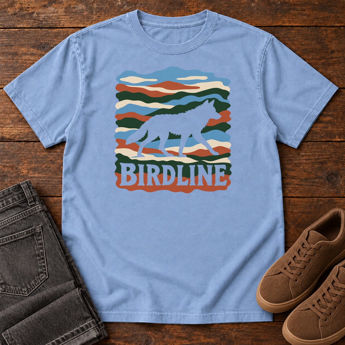 Washed Denim Birdline Wolf Modern T-Shirt with a wolf silhouette over wavy multicolor stripes and Birdline lettering, front flat lay on wood.