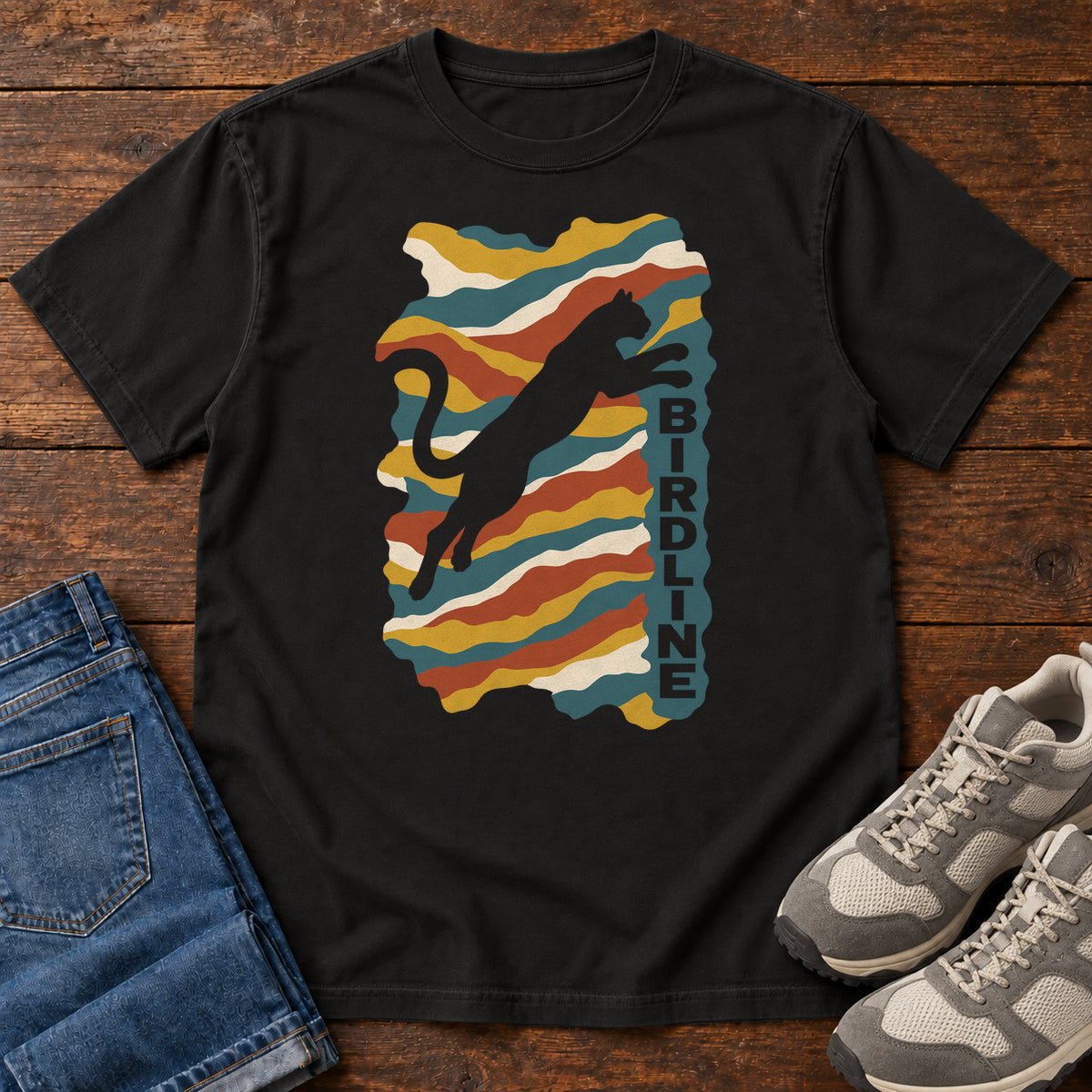 Black Birdline Leaping Cat T-Shirt with a leaping cat silhouette over colorful wavy stripes and vertical Birdline lettering; front flat lay on wood.