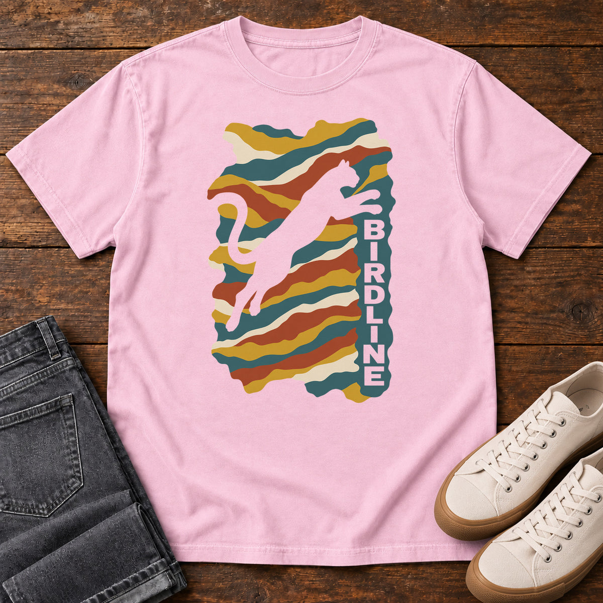 Blossom Birdline Leaping Cat T-Shirt with a leaping cat silhouette over colorful wavy stripes and vertical Birdline lettering; front flat lay on wood.