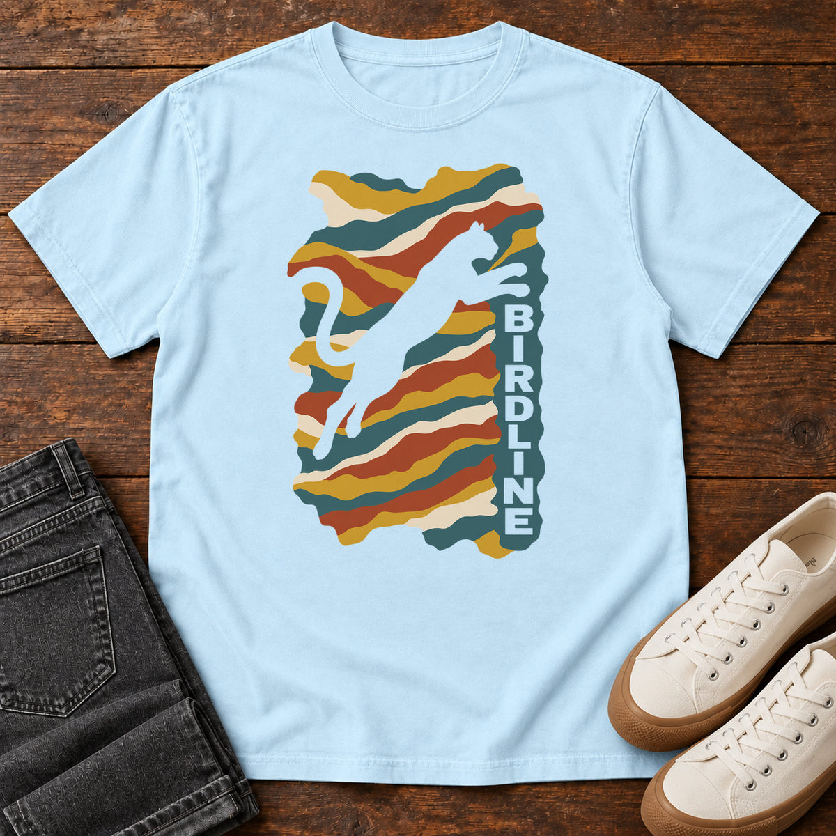 Chambray Birdline Leaping Cat T-Shirt with a leaping cat silhouette over colorful wavy stripes and vertical Birdline lettering; front flat lay on wood.