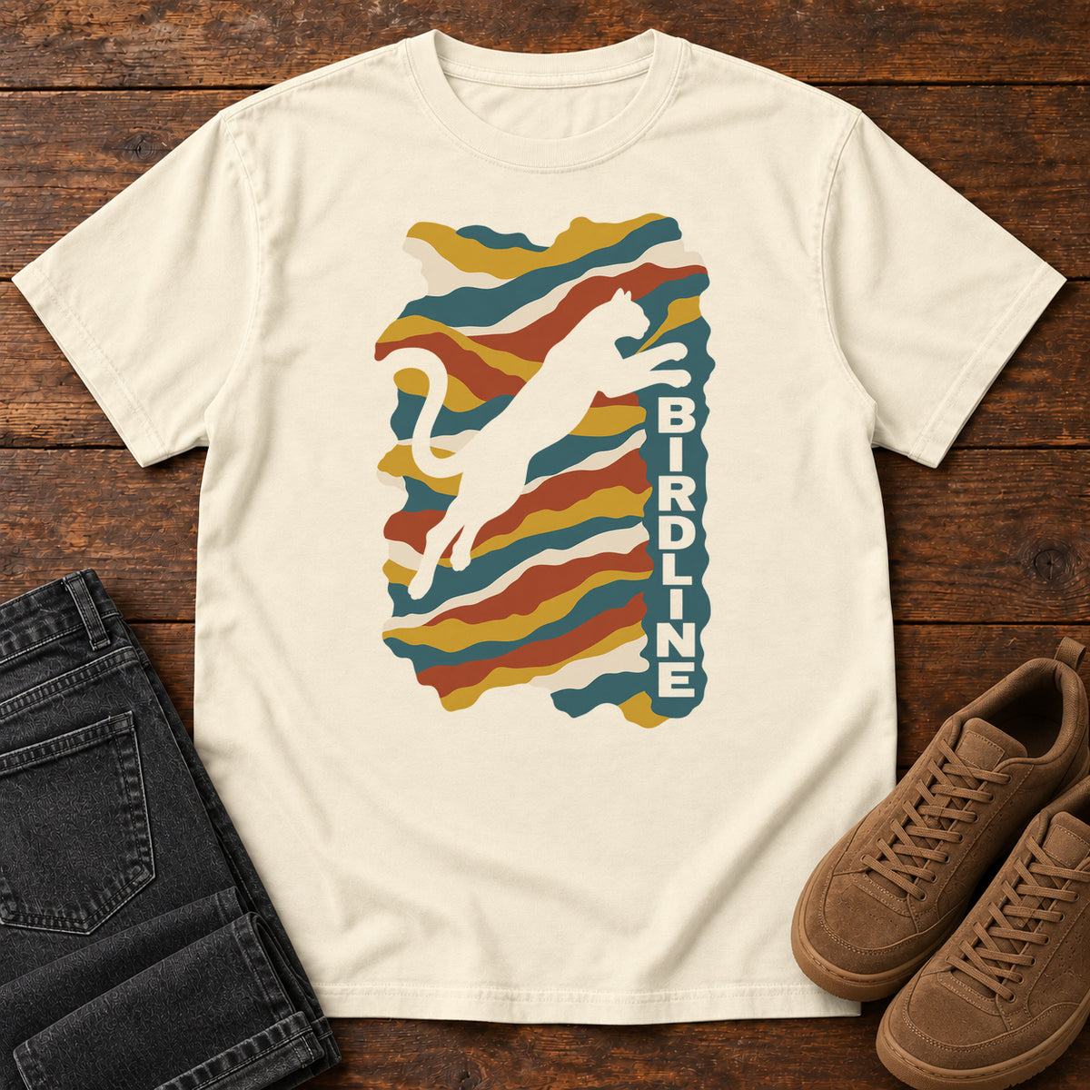 Ivory Birdline Leaping Cat T-Shirt with a leaping cat silhouette over colorful wavy stripes and vertical Birdline lettering; front flat lay on wood.
