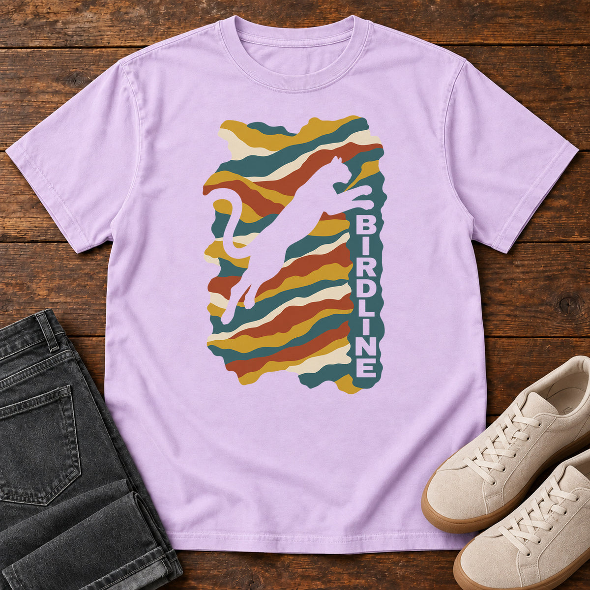 Orchid Birdline Leaping Cat T-Shirt with a leaping cat silhouette over colorful wavy stripes and vertical Birdline lettering; front flat lay on wood.