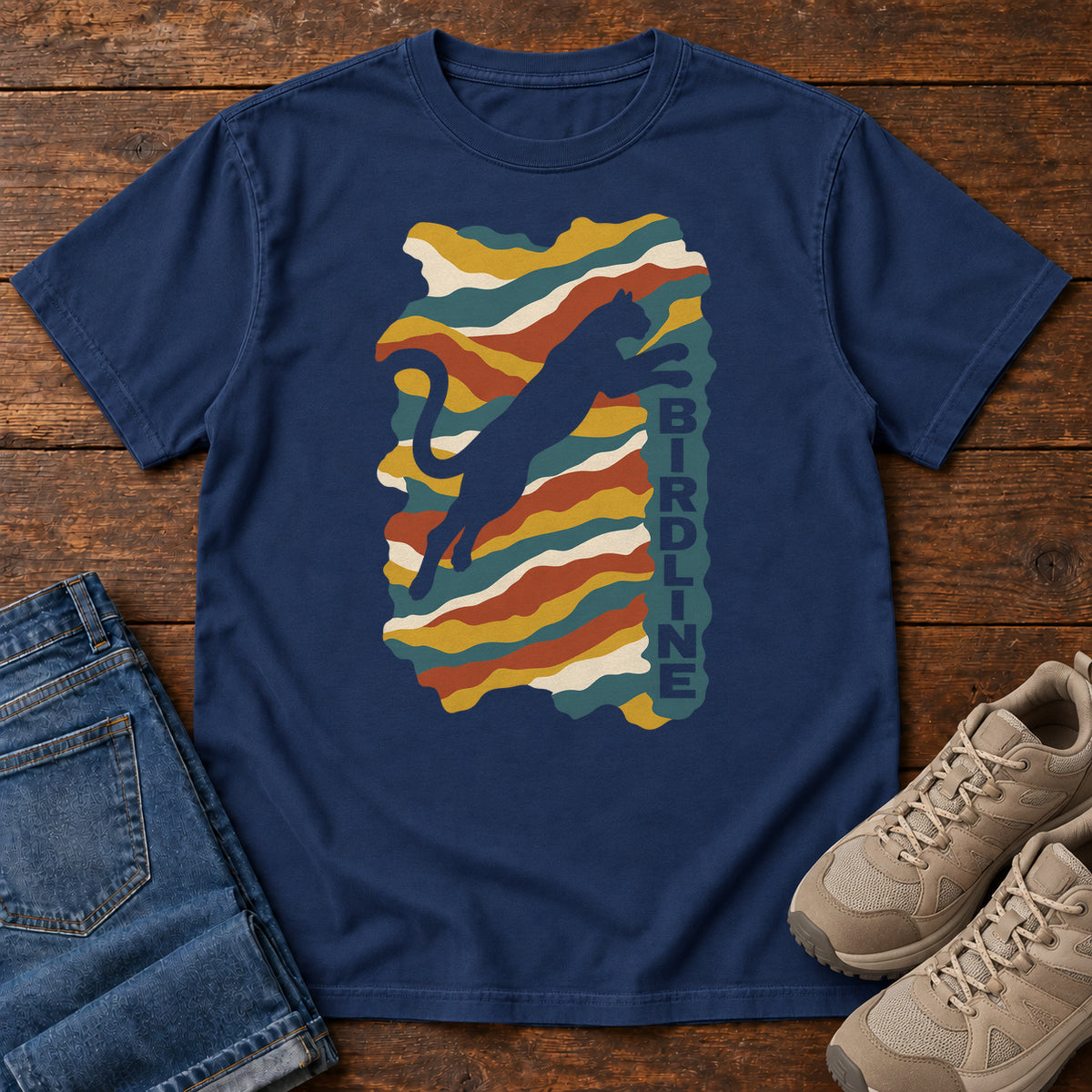 True Navy Birdline Leaping Cat T-Shirt with a leaping cat silhouette over colorful wavy stripes and vertical Birdline lettering; front flat lay on wood.