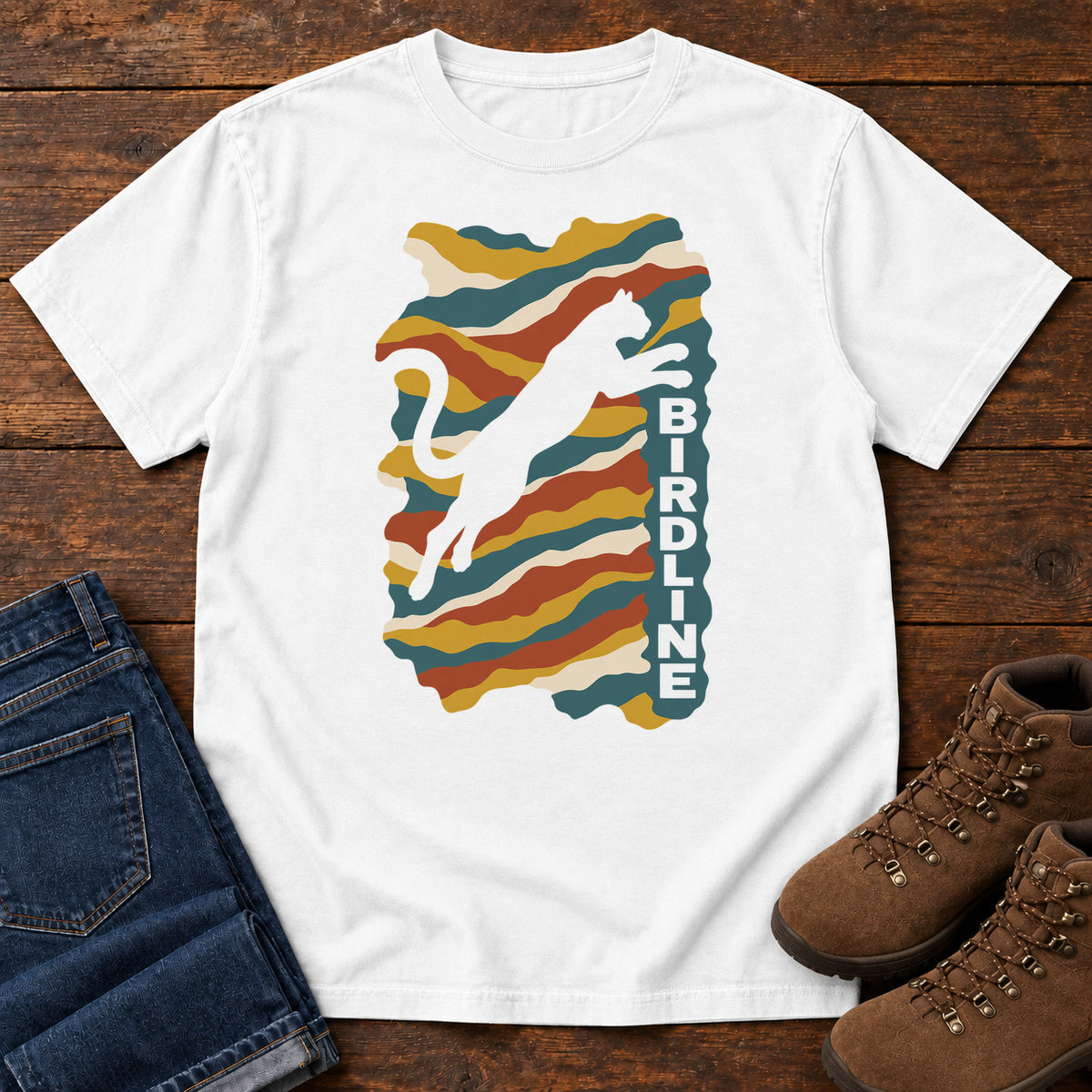 White Birdline Leaping Cat T-Shirt with a leaping cat silhouette over colorful wavy stripes and vertical Birdline lettering; front flat lay on wood.