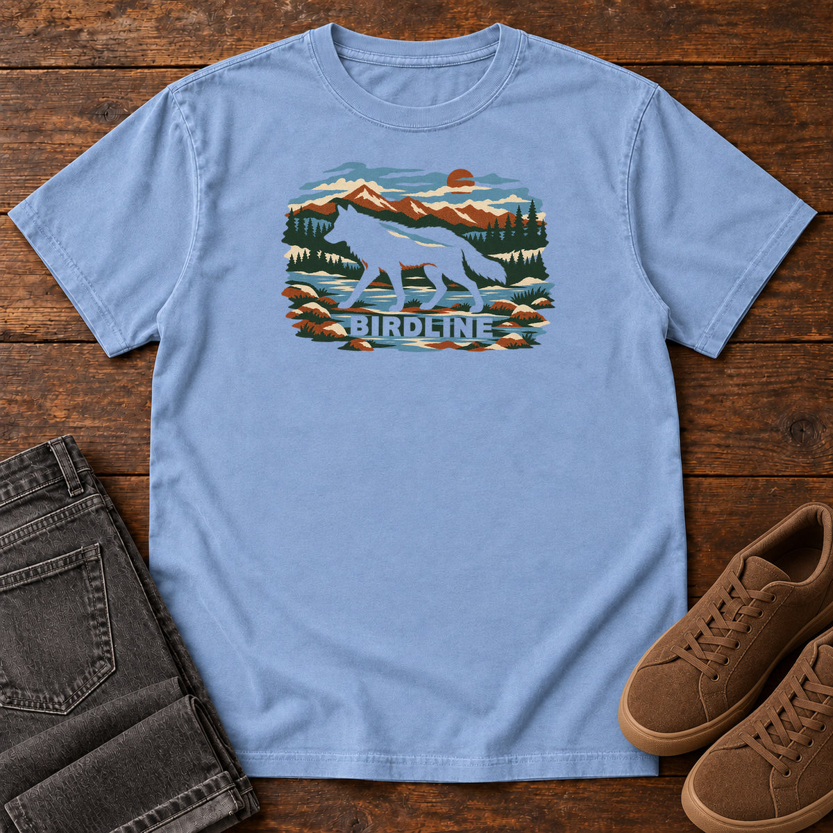 Washed Denim Birdline Wolf Mountain T-Shirt with a wolf silhouette in a mountain, forest and river scene with Birdline lettering, front flat lay on wood.