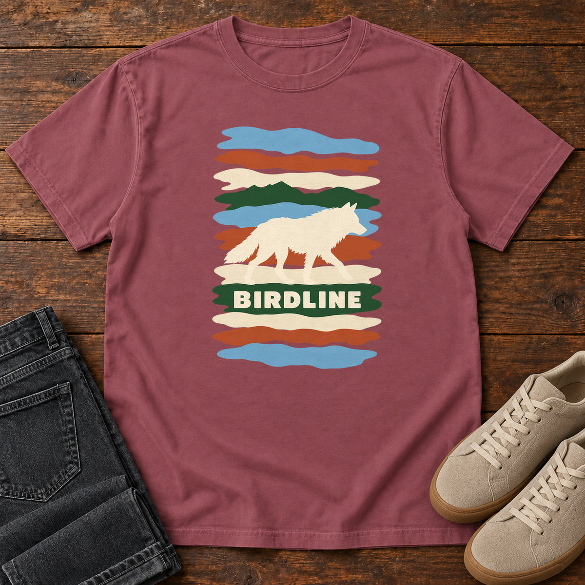 Brick Birdline White Wolf Stripes T-Shirt with a cream wolf silhouette over colorful wavy stripes and Birdline lettering; front flat lay on wood.