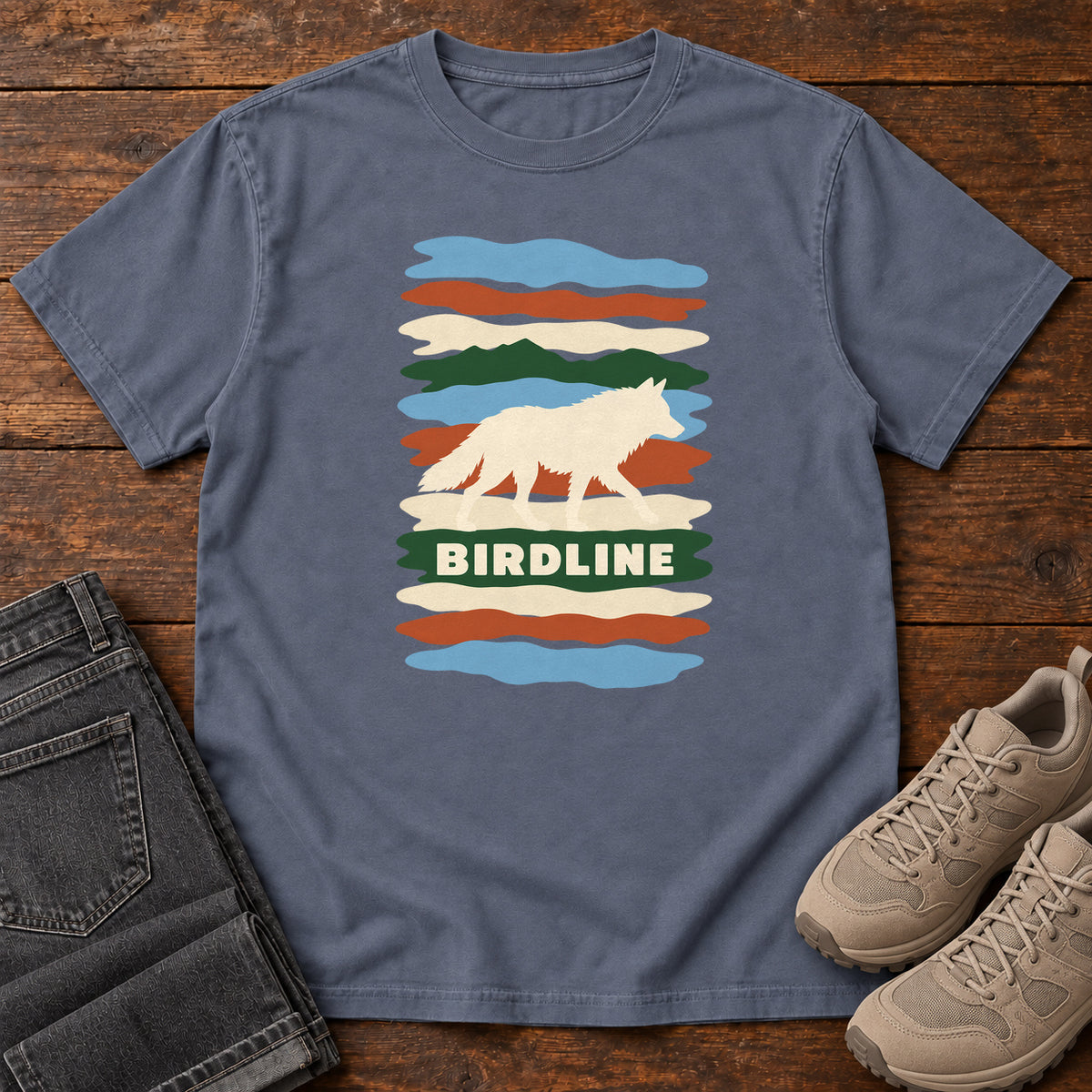 Denim Birdline White Wolf Stripes T-Shirt with a cream wolf silhouette over colorful wavy stripes and Birdline lettering; front flat lay on wood.