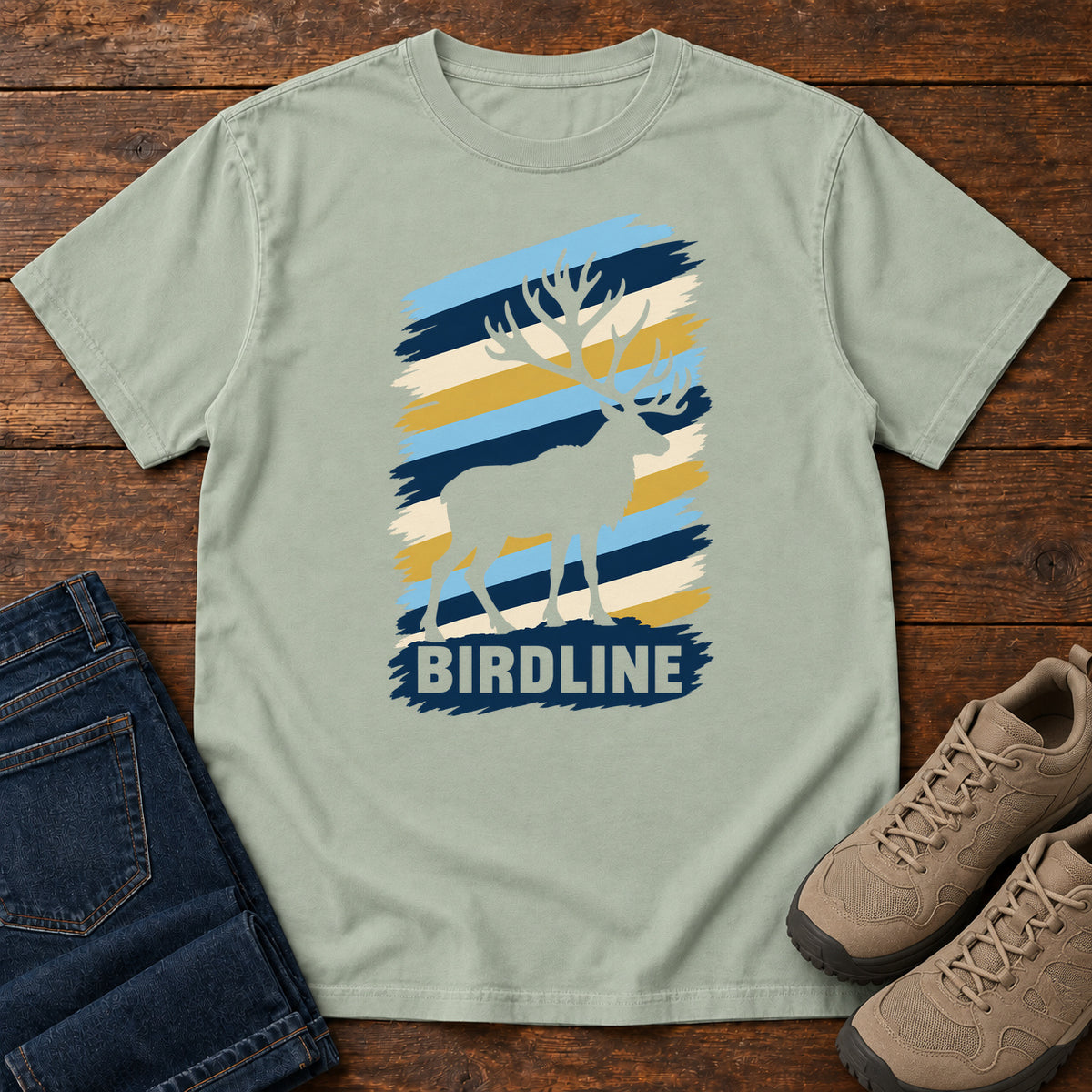 Birdline Elk Stripes T-Shirt in Bay, front laid flat on wood, featuring an elk silhouette against blue, cream and gold brushstroke stripes above BIRDLINE lettering.