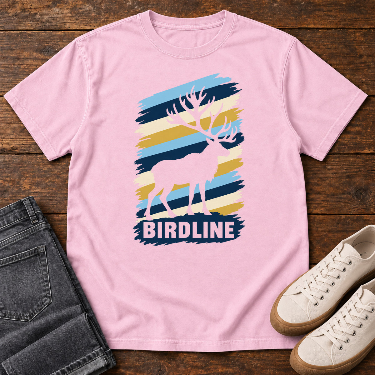 Birdline Elk Stripes T-Shirt in Blossom, front laid flat on wood, featuring an elk silhouette against blue, cream and gold brushstroke stripes above BIRDLINE lettering.