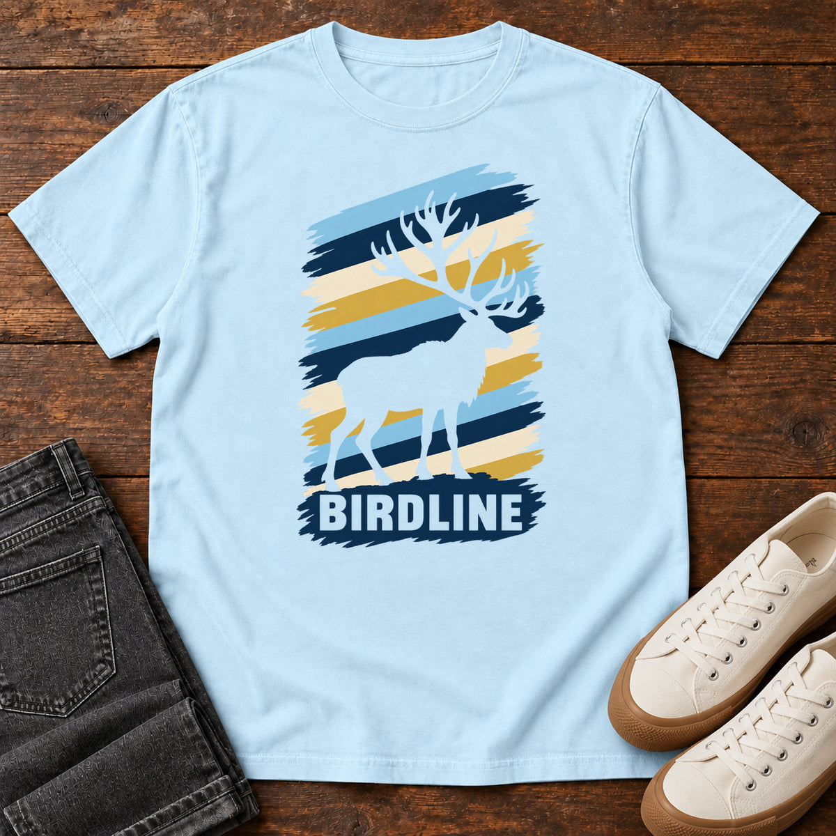 Birdline Elk Stripes T-Shirt in Chambray, front laid flat on wood, featuring an elk silhouette against blue, cream and gold brushstroke stripes above BIRDLINE lettering.