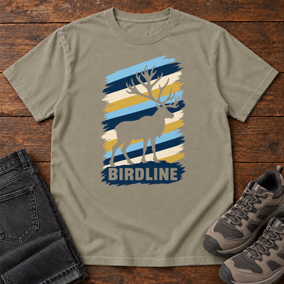 Birdline Elk Stripes T-Shirt in Sandstone, front laid flat on wood, featuring an elk silhouette against blue, cream and gold brushstroke stripes above BIRDLINE lettering.