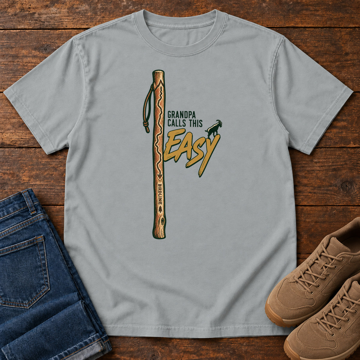 Granite Grandpa Calls This Easy T-Shirt with a wooden hiking stick beside green-and-gold lettering and a small goat silhouette; front flat lay on wood.